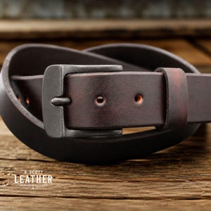 Op de afbeelding: Een donkerbruine leren riem met een matzwarte metalen gesp en een bijpassende leren lus. De riem heeft zichtbare gaatjes en de tekst B. SCOTT LEATHER staat in het wit gedrukt in de linkerbenedenhoek.