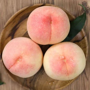 May include: Three ripe peaches with a blush of pink and yellow hues, arranged on a wooden plate. The peaches have a textured surface and a visible seam. A green leaf is visible in the background.