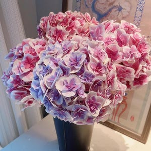 May include: A bouquet of pink and purple hydrangeas in a black metal bucket. The flowers have a gradient of colors, with pink petals fading into purple and white. The arrangement is set against a neutral background.