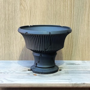 May include: A dark gray ceramic pedestal bowl with a fluted design and visible cracks. The bowl has a wide, slightly flared rim and sits on a pedestal base. The background is a light wood grain.