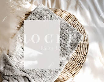 May include: A white card mockup on a woven basket with a gray throw blanket and a fluffy white feather plume.
