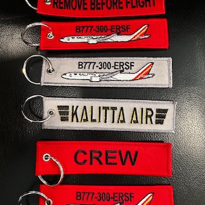 May include: A collection of red and gray embroidered keychains. The keychains feature text such as "REMOVE BEFORE FLIGHT", "CREW", and "KALITTA AIR". Some keychains display an airplane graphic and the text "B777-300-ERSF".