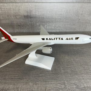 May include: White model airplane of a Boeing 747-8F cargo plane with red and gold accents. The plane is on a white stand and has the text "KALITTA AIR" on the side. The tail number is "N776CK".