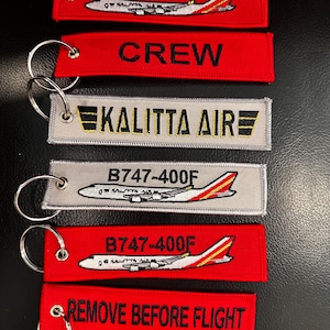 May include: A collection of embroidered keychains in red and silver. The keychains feature text and images of airplanes, including "B747-400F", "CREW", "KALITTA AIR", and "REMOVE BEFORE FLIGHT". Each keychain has a silver keyring.