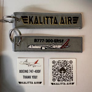 May include: Two gray fabric keychains with metal rings, one with "KALITTA AIR" in gold lettering, the other with a plane illustration and "B777-300-ERSF". Two white square cards with plane graphics and "KALITTA AIR" branding.