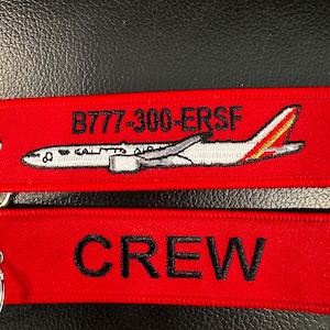 May include: Two red fabric keychains. The top keychain features a white airplane graphic with the text "B777-300-ERSF". The bottom keychain has the word "CREW" embroidered in black.