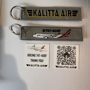May include: Two gray keychains with metal rings. One keychain has the text "KALITTA AIR" embroidered in gold. The other keychain has a Boeing 747-400F airplane design and the text "B747-400F". Two white cards with the "KALITTA AIR" logo and a QR code.