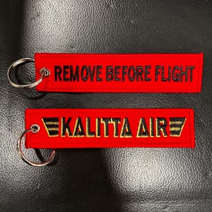 May include: Two red embroidered keychains. One reads "REMOVE BEFORE FLIGHT" in black text. The other reads "KALITTA AIR" in gold and black text. Both have silver key rings.