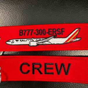 May include: Two red fabric keychains. The top keychain features a white airplane graphic with the text "B777-300-ERSF". The bottom keychain has the word "CREW" embroidered in black.