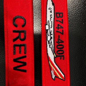 May include: Two red fabric keychains. One keychain says "CREW" in black letters. The other keychain has a white airplane graphic with the text "B747-400F" and "QW KALITTA AIR" in black.