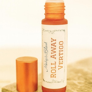 Puede incluir: Una botella roll-on naranja con el texto "Roll Away Vertigo" y "Mulye's Blend" en una etiqueta blanca. La botella tiene una bola rodante plateada y una tapa naranja separada. El producto se exhibe sobre una superficie de mármol.