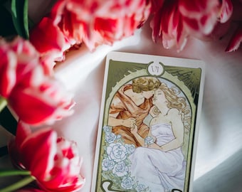 Puede incluir: Una carta del tarot con un marco dorado y un fondo verde. La carta representa a una pareja abrazándose, con la mujer vestida con un vestido blanco y el hombre con una túnica marrón. La carta se titula "Los Amantes" y tiene el número romano VI en la esquina superior derecha.