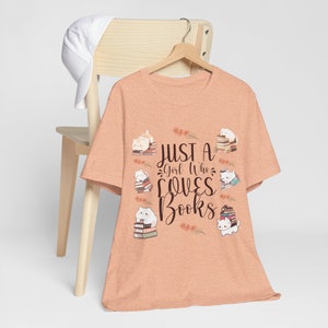May include: A peach-colored t-shirt with the text "Just a Girl Who Loves Books" and illustrations of cats sitting on stacks of books.