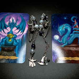 May include: A pair of tarot card bookmarks with silver-tone charms. The bookmarks are between two tarot cards. One card shows a blue dragon holding a cup, labeled "Ace of Cups." The other card depicts a blue dragon with a glowing orb, labeled "The Hermit."