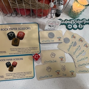 Puede incluir: Un juego de cartas de Piedra Papel Tijeras con un dado y tarjetas para rascar. Las cartas son amarillas con un borde marrón y muestran una imagen de una mano haciendo un gesto de piedra, papel o tijeras. El texto en las cartas dice "Windmill Ridge Budgets".