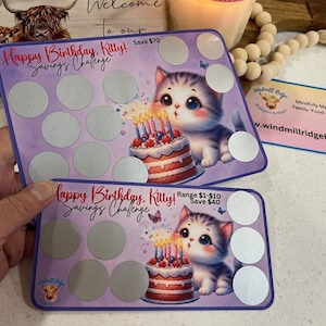 以下が含まれることがあります： 子猫と誕生日ケーキが描かれた、紫と白のカード2枚。カードには銀色のコーティングが施された円形のグリッドがあります。カードのテキストは「Happy Birthday, Killy! Savings Challenge」と「Happy Birthday, Killy! Savings Challenge」です。
