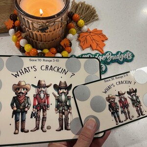 Può includere: Due pannelli bianchi con il testo "What's Crackin?" e un intervallo di numeri. I pannelli hanno un design con tre figure di schiaccianoci con cappelli da cowboy. I pannelli sono su una superficie bianca con una candela e decorazioni autunnali sullo sfondo.