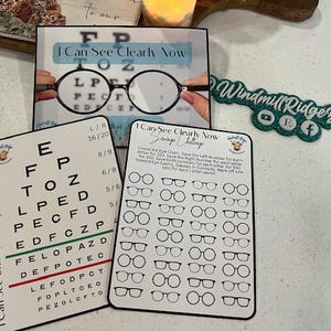 May include: Two white cards with black text and illustrations. The top card has a large eye chart with the text "I Can See Clearly Now" at the top. The bottom card has a smaller eye chart with the text "I Can See Clearly Now Savings Challenge" at the top. The card has a series of black and white illustrations of eyeglasses.