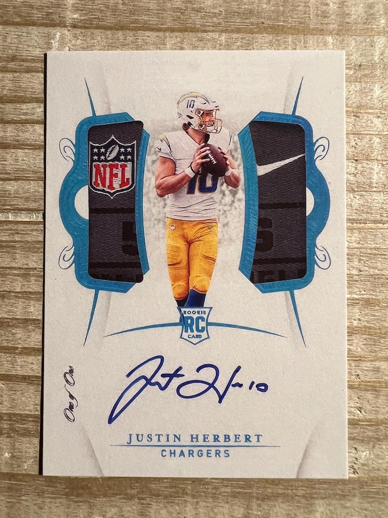 Op de afbeelding: Een witte en blauwe voetbalkaart met Justin Herbert, quarterback van de Los Angeles Chargers. De kaart heeft aan elke kant een blauw en zwart jersey-patch. De kaart is gesigneerd met "Justin Herbert 10".