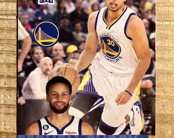 Könnte beinhalten: Basketball-Sammelkarte mit Stephen Curry von den Golden State Warriors. Die Karte enthält ein Rookie Card-Logo, ein Foto von Curry in Aktion und ein kleineres Porträt. Das Teamlogo und der Spielername sind ebenfalls sichtbar.