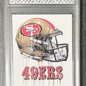 May include: A graded rookie card of Brock Purdy, the quarterback for the San Francisco 49ers. The card features a gold and red illustration of a football helmet with the 49ers logo. The text "49ERS" is printed in red below the helmet. The text "Brock Purdy #13" and "2022 Rookie Card" is printed below the illustration.