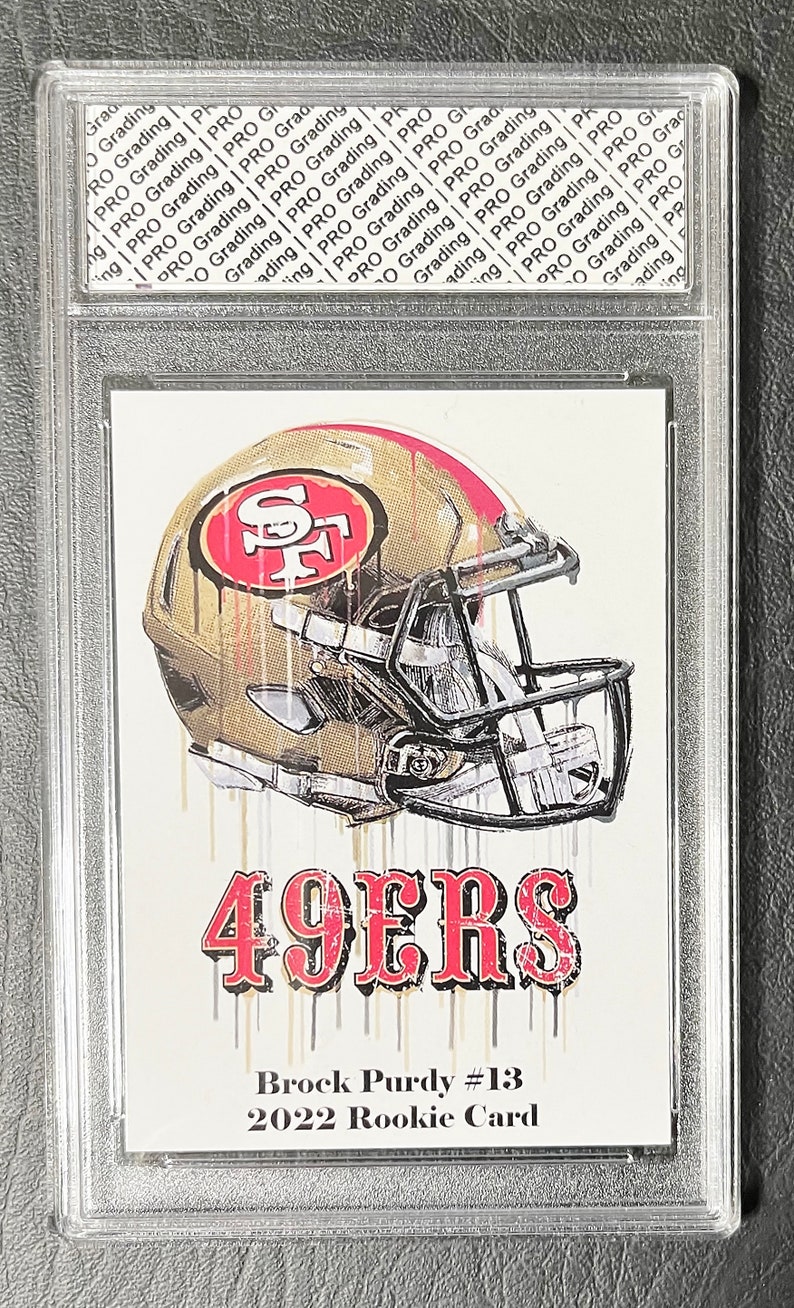 May include: A graded rookie card of Brock Purdy, the quarterback for the San Francisco 49ers. The card features a gold and red illustration of a football helmet with the 49ers logo. The text "49ERS" is printed in red below the helmet. The text "Brock Purdy #13" and "2022 Rookie Card" is printed below the illustration.