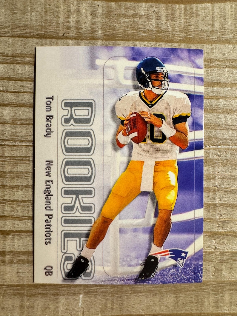 May include: A rookie card featuring Tom Brady, quarterback for the New England Patriots, in a white jersey with the number 10. The card has a blue and white background with the word "Rookies" in gray letters.