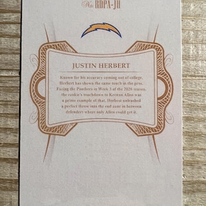 Op de afbeelding: Een witte kaart met een bruine rand en een gouden ontwerp. De kaart heeft een blauw bliksemlogo en de tekst "Justin Herbert" in een bruin lettertype. De tekst "No. RDPA-JH" staat in de rechterbovenhoek van de kaart.