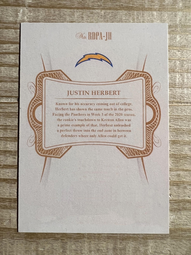 Op de afbeelding: Een witte kaart met een bruine rand en een gouden ontwerp. De kaart heeft een blauw bliksemlogo en de tekst "Justin Herbert" in een bruin lettertype. De tekst "No. RDPA-JH" staat in de rechterbovenhoek van de kaart.