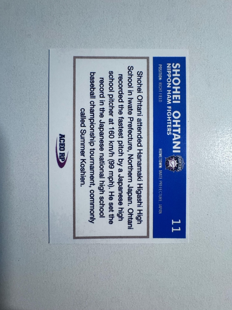 May include: A baseball card for Shohei Ohtani of the Nippon Ham Fighters. The card is blue and white, displaying the player's name, team, position, and hometown. Text details Ohtani's high school record, including a 160 km/h pitch.
