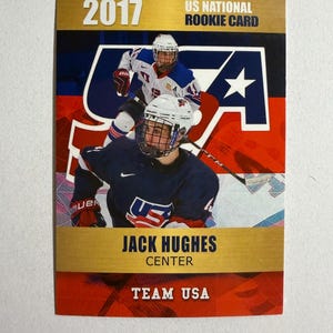 May include: A 2017 US National Rookie Card featuring Jack Hughes, Center, Team USA. The card has a gold border and a background with red, white, and blue colors. The card displays the USA logo and images of hockey players in action.