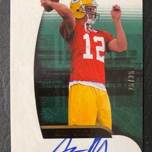 May include: A Green Bay Packers football trading card showing a player in a yellow helmet and red jersey throwing a football. The card has an autograph and the text "Aaron Rodgers" and "Green Bay Packers". The card is numbered 25/25.