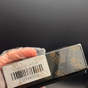 Pode incluir: Uma caixa retangular preta com detalhes dourados e texto árabe. Uma etiqueta branca com um código de barras e o número 23004003 é visível. A caixa é segurada por uma mão enluvada.