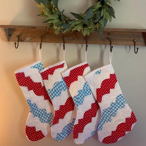 Puede incluir: Cuatro calcetines navideños acolchados con diseños rojos y azules cuelgan de un estante de madera. Una corona verde con pequeñas luces está encima del estante, creando una exhibición festiva.