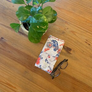 May include: A white quilted glasses case with a floral and koi fish pattern, next to a pair of eyeglasses. A potted plant with green leaves is in the background. The case has a small label with a logo.