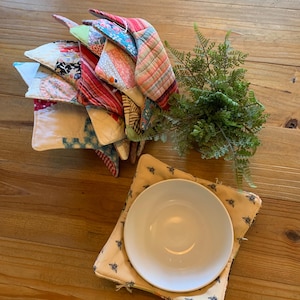 May include: A stack of colorful, quilted pot holders with various patterns and colors, including red, blue, and floral designs. A white plate rests on a square pot holder with a bee pattern. A sprig of greenery is also present.
