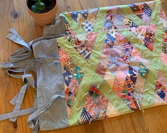 May include: A handmade quilt with a patchwork design featuring various fabric patterns and colors, including red, blue, and green. The quilt is paired with a gray apron with long ties, all resting on a wooden surface.