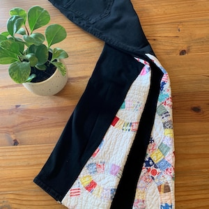 May include: Black denim jeans with a colorful patchwork quilt design on the outer seams. The quilt features a variety of fabric patterns and colors, including red, blue, yellow, and green. The jeans are laid out on a wooden surface.