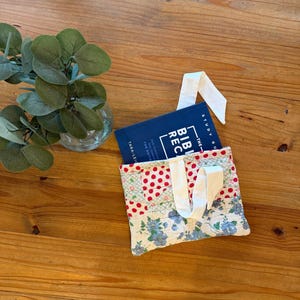 May include: A quilted fabric book sleeve featuring a patchwork design of red polka dots and blue floral patterns on a white background, with white ribbon ties. A blue book titled The Bible Recap Study Guide is tucked inside the sleeve.