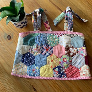 May include: A handmade patchwork bag with a pink gingham trim. The bag features a variety of colorful fabric patches in hexagonal shapes. Two fabric straps are attached to the bag. A potted plant with green leaves sits to the left.