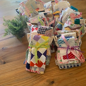 May include: A collection of colorful, patchwork quilt squares, some bundled with ribbons. The squares feature various patterns and colors, including red, blue, and yellow. A small potted plant sits to the left on a wooden table.