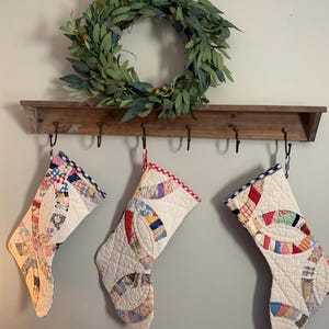 Puede incluir: Tres calcetines navideños acolchados con diseños de patchwork coloridos cuelgan de una repisa de madera con ganchos. Una corona verde con bayas se encuentra encima, creando un ambiente festivo.