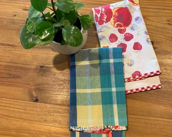 Könnte beinhalten: Draufsicht auf zwei gefaltete Stoffservietten und eine Topfpflanze. Eine Serviette hat ein blaues, gelbes und grünes Karomuster. Die andere Serviette hat einen weißen Hintergrund mit roten Erdbeeren und Blumen. Die Pflanze hat grüne Blätter.