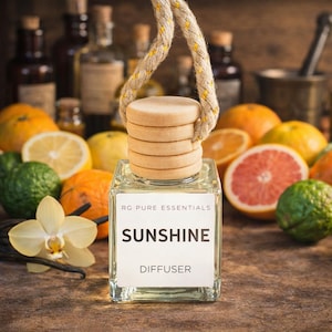 May include: A clear glass diffuser with a wooden cap and a hanging rope. The label reads "SUNSHINE DIFFUSER" and "RG PURE ESSENTIALS". The background features citrus fruits like oranges, lemons, and grapefruit, and a vanilla orchid.