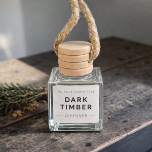 May include: A square glass diffuser bottle with a wooden cap and a woven rope. The label reads "RG PURE ESSENTIALS DARK TIMBER DIFFUSER". The bottle is on a wooden surface.