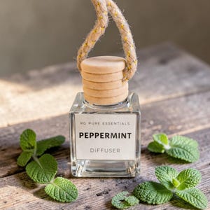 May include: A clear, square glass diffuser bottle with a wooden cap and a rope handle. The label reads "RG Pure Essentials" and "Peppermint Diffuser." Fresh mint leaves are scattered around the bottle on a wooden surface.