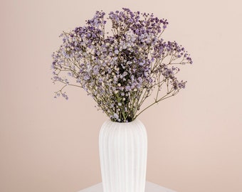 May include: A white ceramic vase with a textured surface holds a bouquet of purple and white baby's breath flowers. The vase is sitting on a white pedestal.