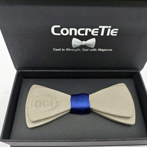 Pode incluir: Uma gravata borboleta de concreto com uma fita azul, apresentada em uma caixa preta. A tampa da caixa exibe o texto "ConcreTie" e "Cast in Strength, Tied with Elegance". A gravata borboleta tem um design único e em camadas.