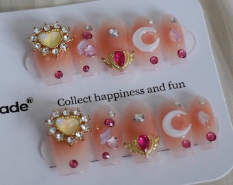May include: A set of ten pink press-on nails with various gold and white embellishments, including hearts, moons, and wings. The nails are decorated with rhinestones and the text "Collect happiness and fun" is printed on the white background.