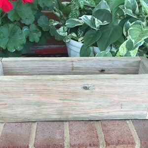 May include: A rectangular wooden planter box made from weathered, light-colored wood. The box has a simple, open design, perfect for holding plants. The wood has a natural, rustic appearance, with visible grain and knots. The planter is sitting on a brick surface.